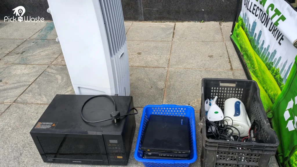 Old electronics collected during the drive, including a Samsung microwave, an air cooler, a laptop in a blue basket, and small household appliances in a crate.